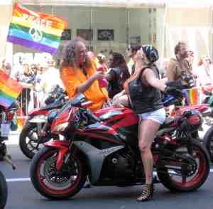 Dyke on Bike getting spray misted by Tiny Tim lookalike.